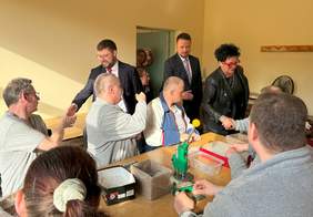 Pokaż zdjęcie: Przywitanie sie przedstawicieli PFRON w trakcie kolejnych zajęć w MZAZ w Dobrej. Osoby siedzące przy stole i reprezentacja PFRON stoi w drzwiach.