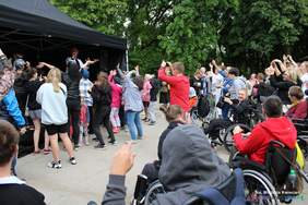 Pokaż zdjęcie: Występ zespołu disco polo, przed scena stoi grupa osób tańczących do muzyki, część osób siedzi na wózkach inwalidzkich