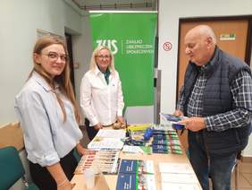 Pokaż zdjęcie: Wokół stołu stoją dwie kobiety i jeden mężczyzna. Rozmawiają ze sobą i uśmiechają się. Na stole leżą różne informatory, poradniki, instruktarze, gadżety. 