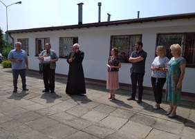 Pokaż zdjęcie: Siedem osób - kobiety i mężczyźni stoją na tle budynku. Słuchają przemawiającego księdza 