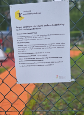 Pokaż zdjęcie: Na ogrodzeniu wisi tablica informacyjna, na której zamieszczone są informacje na temat dofinansowania przez PFRON budowy placu zabaw w ramach programu Dostępna Przestrzeń Publiczna. 