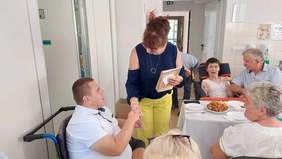 Pokaż zdjęcie: Kobieta w granatowej bluzce i zielonych spodniach ściska rękę mężczyźnie siedzącemu na wózku inwalidzkim. 