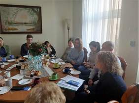 Pokaż zdjęcie: pomieszczenie w środku budynku grupa osób siedzi przy okrągłym stole, na którym umieszczone są materiały o projekcie powrót do zdrowia powrót do pracy. Prowadzona jest dyskusja z uczestnikami spotkania