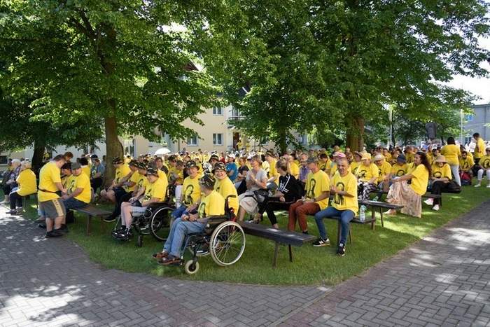 Pokaż zdjęcie: Grupa niepełnosprawnych podczas Pielgrzymki w żółtych koszulkach siedzą na ławkach w plenerze