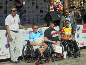 Pokaż zdjęcie: Pamiątkowe zjęcie pierwszej trójki zwycięzców w kategorii "wózkarzy hanbike" Mężczyzn. Za sportowcami stoji Dyrektor Oddziału Pomorskiego PFRON.