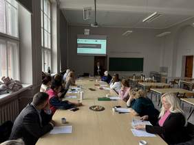 Pokaż zdjęcie: Sala wykładowa, przy długim drewnianym stole siedzi kilkanaście osób, na końcu stołu mężczyzna w okularach i marynarce mówi do siedzących. Za nim telebim z prezentacją.