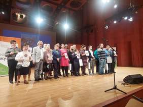 Pokaż zdjęcie: na zdjęciu scena sali koncertowej Akademii Muzycznej na której stoi grupa osób - laureatów konkursu plastycznego oraz członkowie komisji konkursowej, laureaci trzymają nagrody, dyplomy i statuetki z logo PFRON