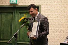 Pokaż zdjęcie: Przed mikrofonem stoi lekko schylony młody mężczyzna w szarym garniturze w ciemna delikatna kratę, w białej koszuli z czarnym krawatem. W lewej ręce trzyma dyplom oraz statuetkę lodołamaczy a w prawej bukiet kwiatów.