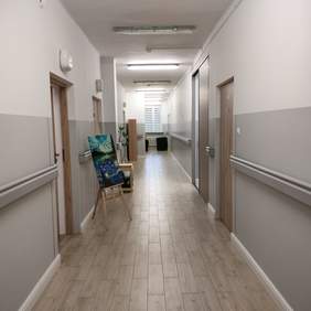 Pokaż zdjęcie: Zdjęcie korytarza pomalowanego na biało szary kolor z lamperią. Po obydwu stronach zamontowane są poręcze. Drzwi do pokoi w kolorze jasnego drewna, na podłodze gres szkliwiony imitujący drewniane panele