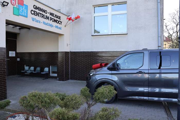 Pokaż zdjęcie: Szary bus z czerwoną kokardą na masce stoi zaparkowany przed wejściem do budynku, na którym widnieje napis: „Gminno-Miejskie Centrum Pomocy Wiara-Nadzieja-Miłość”. Po lewej stronie znajdują się trzy krzesła przymocowane do ściany budynku.