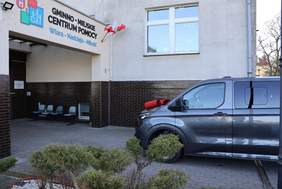 Pokaż zdjęcie: Szary bus z czerwoną kokardą na masce stoi zaparkowany przed wejściem do budynku, na którym widnieje napis: „Gminno-Miejskie Centrum Pomocy Wiara-Nadzieja-Miłość”. Po lewej stronie znajdują się trzy krzesła przymocowane do ściany budynku.