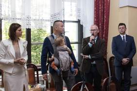 Pokaż zdjęcie: W jasnym pomieszczeniu, z oknami w tle, cztery stoją obok siebie. Kobieta w jasnym kostiumie stoi po lewej, obok niej mężczyzna w marynarce z dzieckiem w nosidełku. Centralnie, mężczyzna w okularach przemawia do mikrofonu, gestykulując. Po prawej stoi mężczyzna ubrany w garnitur.