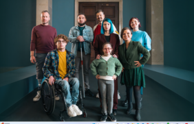 Pokaż zdjęcie: Na ciemnym tle stoi grupa osób z niepełnosprawnościami. Wszyscy patrzą w obiektyw aparatu, a niektórzy się uśmiechają. W tle widać ściany, filary i drzwi. 