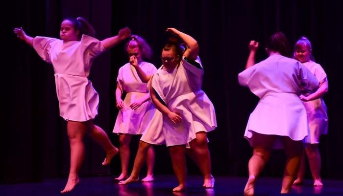 Pokaż zdjęcie: Na scenie występuje 6 dziewczynek tak samo ubranych w jasne sukienki, dziewczyny tańczą.