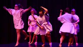 Pokaż zdjęcie: Na scenie występuje 6 dziewczynek tak samo ubranych w jasne sukienki, dziewczyny tańczą.