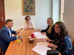 Pokaż zdjęcie: Przy stole siedzą trzy kobiety i mężczyzna. Kobiety po prawej uśmiechają się do zdjęcia i podpisują dokumenty. Mężczyzna po lewej ubrany w niebieską marynarkę czyta dokumenty które trzyma w ręku.