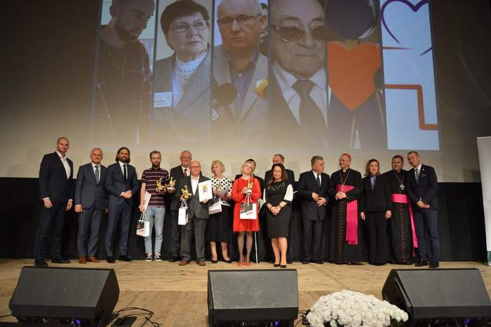 Pokaż zdjęcie: Widok na scenę w Domu Kultury w Świebodzinie, laureaci nagrody stoją obok siebie, na tle prezentacji, są w garniturach, strojach galowych, jest jasno i dużo fleszy