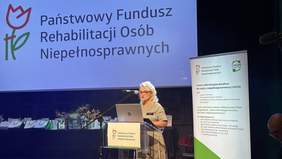 Pokaż zdjęcie: Ubrana w sukienkę kobieta w trakcie przemówienia na tle logo PFRON i baneru CIDON