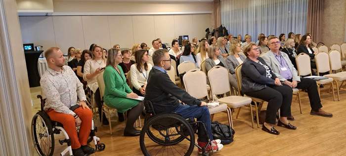 Na sali konferencyjnej siedzą uczestnicy konferencji, organizowanej przez PFRON. Po lewej stronie widzocznisą dwaj meżczyni na wózkach inwalidzkich.