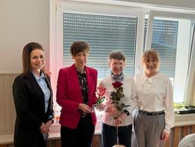 Zdjęcie grupowe czterech kobiet stojących obok siebie w pomieszczeniu, na tle dużego okna z roletą. Od lewej: kobieta w czarnym garniturze i jasnoniebieskiej koszuli, z długimi brązowymi włosami. Druga kobieta ma krótkie ciemne włosy i czerwony żakiet i trzyma w ręku statuetkę. Trzecia kobieta ma krótkie włosy z grzywką, białą bluzkę i trzyma w rękach czerwoną różę. Czwarta kobieta ma jasnobrązowe włosy z grzywką, białą bluzkę i szare spodnie. 