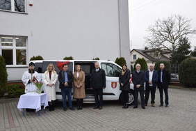 Pokaż zdjęcie: Na tle białego busa marki FORD stoi grupa osób pozująca do pamiątkowego zdjęcia. 