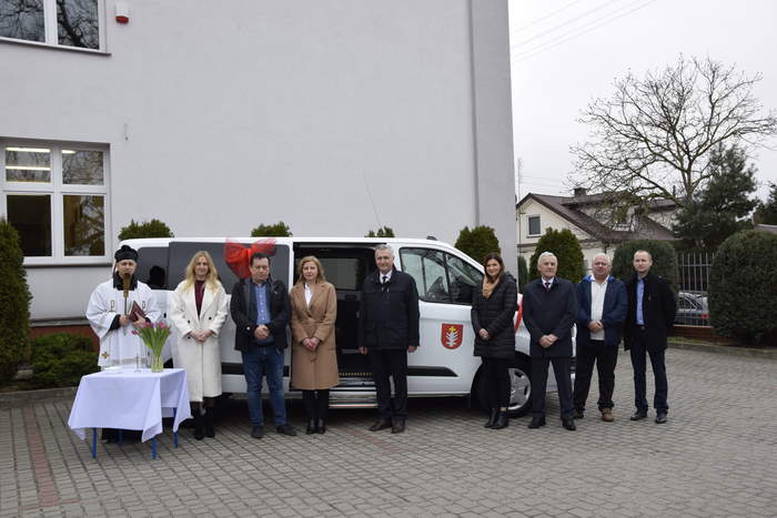 Pokaż zdjęcie: Na tle białego busa marki FORD stoi grupa osób pozująca do pamiątkowego zdjęcia. 