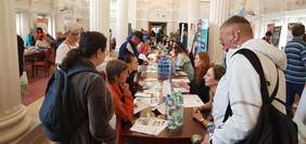 W przestronnej sali z białymi kolumnami uczestnicy wydarzenia siedzą przy długim stole bądź przemieszczają się