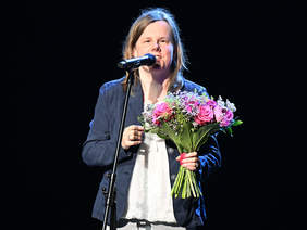 Pokaż zdjęcie: Osoba stojąca na scenie przy mikrofonie, trzyma bukiet różowych i fioletowych kwiatów. Ubrana w niebieską marynarkę i jasną bluzkę.
