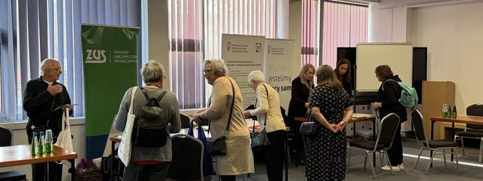 Pokaż zdjęcie: Grupka osób w sali konferencyjnej rozmawia przy stolikach informacyjnych z materiałami ZUS i innych instytucji; jasne pomieszczenie z dużymi oknami, krzesłami i stołami ustawionymi do obsługi uczestników.