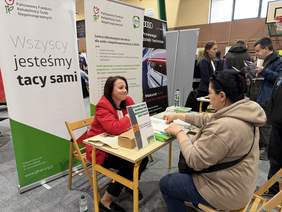 Pokaż zdjęcie: Stoisko PFRON, CIDON podczas wydarzenia w hali sportowej. Kobieta w czerwonej marynarce rozmawia z uczestniczką siedzącą przy stoliku.