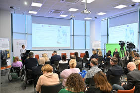 Pokaż zdjęcie: sala konferencyjna (ujęcie z tyłu sali), grupa osób mężczyźni i kobiety, siedzą na krzesłach w dwóch rzędach