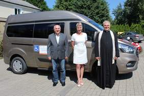 Pokaż zdjęcie: Na tle busa stoiod lewej mężczyzna ubrany w szarą marynarkę, kobieta ubrana w białą sukienkę oraz ksiądz 