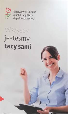 Pokaż zdjęcie: Plakat PFRON z napisem "Wszyscy jesteśmy tacy sami" i z logo funduszu, na wózku inwalidzkim siedzi uśmiechnięta brunetka, w niebieskiej koszuli, z długopisem w dłoni