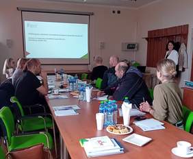 Pokaż zdjęcie: Grupa osób siedzi przy długim stole konferencyjnym, uczestnicząc w prezentacji. Na ekranie wyświetlany jest slajd zawierający informacje o dotacjach ze środków PFRON na rozpoczęcie działalności gospodarczej oraz o refundacji składek ZUS. Na stole znajdują się dokumenty, butelki z wodą, filiżanki oraz broszury informacyjne. Prezentację prowadzi kobieta stojąca obok ekranu.