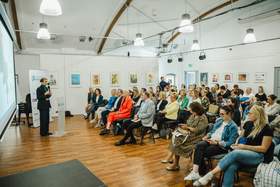 Sala konferencyjna z drewnianą podłogą i białymi ścianami z zawieszonymi ilustracjami dzieci. Kobieta w czarnym garniturze przemawia przy mównicy z mikrofonem do siedzącej publiczności. 