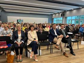 Pokaż zdjęcie: W auli ustawione są rzędy krzeseł, na których siedzą ludzie. Trwa konferencja dotycząca zdrowia psychicznego dzieci i młodzieży. 