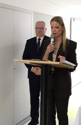 Pokaż zdjęcie: Na zdjęciu kobieta w eleganckim garniturze prowadzi przemówienie, stojąc przy mównicy, na której leży otwarta książka. W dłoni trzyma mikrofon. Obok niej stoi mężczyzna w ciemnym garniturze. W tle widać część nowoczesnego pomieszczenia.