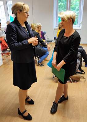 Pokaż zdjęcie: Dwie kobiety elegancko ubrane stoją na środku sali i rozmawiają. 