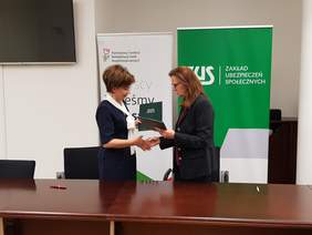 Pokaż zdjęcie: Dwie elegancko ubrane kobiety podają sobie ręce. Wymieniają się też teczkami z dokumentami.