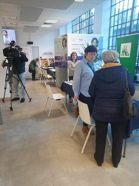 Pokaż zdjęcie: W sali znajdują się różne osoby. Dwie panie stoją i rozmawiają ze sobą, inne osoby siedzą przy stoiskach. W tle widać operatora z kamerą telewizyjną. 