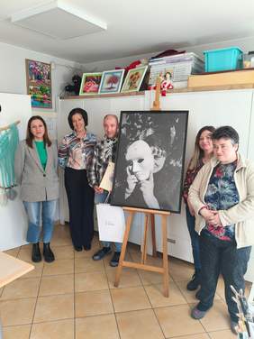 Pokaż zdjęcie: Grupa osób w pracowni artystycznej ogląda dużą fotografię ustawioną na sztaludze. Jedna z osób prezentuje dyplom i torbę z nagrodą. W tle wiszą liczne kolorowe prace plastyczne i wyposażenie pracowni.