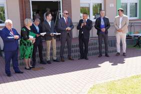 Pokaż zdjęcie: Osiem osób stoi przed budynkiem, świeci słońce, są ubrani elegancko. Jeden z mężczyzn trzyma mikrofon i przemawia.