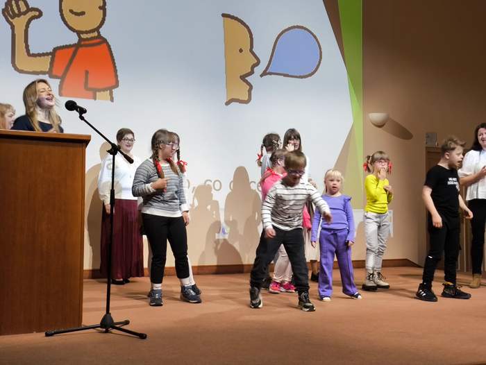 Pokaż zdjęcie: Scena na której tańczą kolorowo ubrane przedszkolaki. W tle piktogramy z mową.