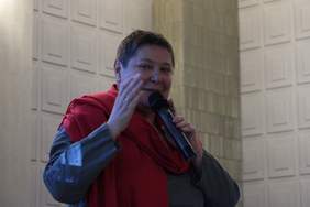 Pokaż zdjęcie: Uśmiechnięta kobieta w średnim wieku, krótko ostrzyżona trzyma w ręku mikrofon i spogląda w obiektyw. Ubrana jest w niebieską bluzkę, na szyi ma czerwony szal