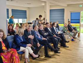 Widok na widownię siedzącą w rzędzie na niebieskich krzesłach w jasnej, nowoczesnej sali szkoleniowej z drewnopodobną podłogą. W pierwszym rzędzie siedzi kilkanaście osób – głównie mężczyźni w garniturach oraz kobiety w eleganckich strojach. Po lewej stronie starszy mężczyzna w marynarce i krawacie rozmawia z siedzącą obok kobietą w granatowej, błyszczącej marynarce. Dalej w rzędzie siedzą mężczyźni znani z poprzednich zdjęć (m.in. w jasnoniebieskiej marynarce i ciemnych garniturach). W głębi sali, za filarem, widać stojącego kamerzystę z gimbalem oraz inne osoby obserwujące spotkanie. Okna w tle zasłonięte są niebieskimi roletami rzymskimi.