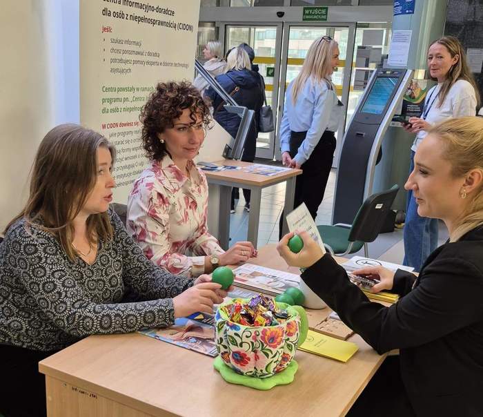 Pokaż zdjęcie: Przy biurku siedzą i rozmawiają trzy kobiety. Jedna z nich trzyma zieloną piłkę antystresową w ręku. W tle stoi baner z logo CIDON.