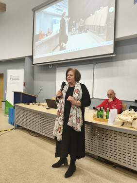 Kobieta w czarnej sukience i kolorowym szalu, stoi, w ręce trzyma mikrofon