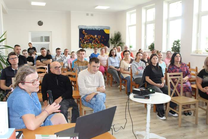 Pokaż zdjęcie: Pracownik PFRON w trakcie wystąpienia na tle zgromadzonych uczestników spotkania, którzy siedzą na krzesłach