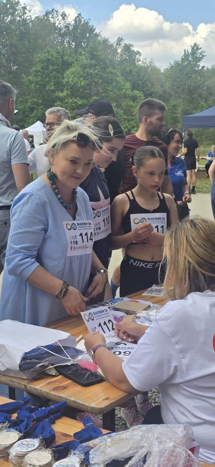 Pokaż zdjęcie: Kobieta ubrana na niebiesko z plastronem uczestnika biegu stoi przy stoliku rejestracji, a obok niej dziewczynka i inne osoby