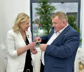 Pokaż zdjęcie: Kobieta w białej marynarce i mężczyzna w garniturze składają sobie życzenia i dzielą się opłatkiem.